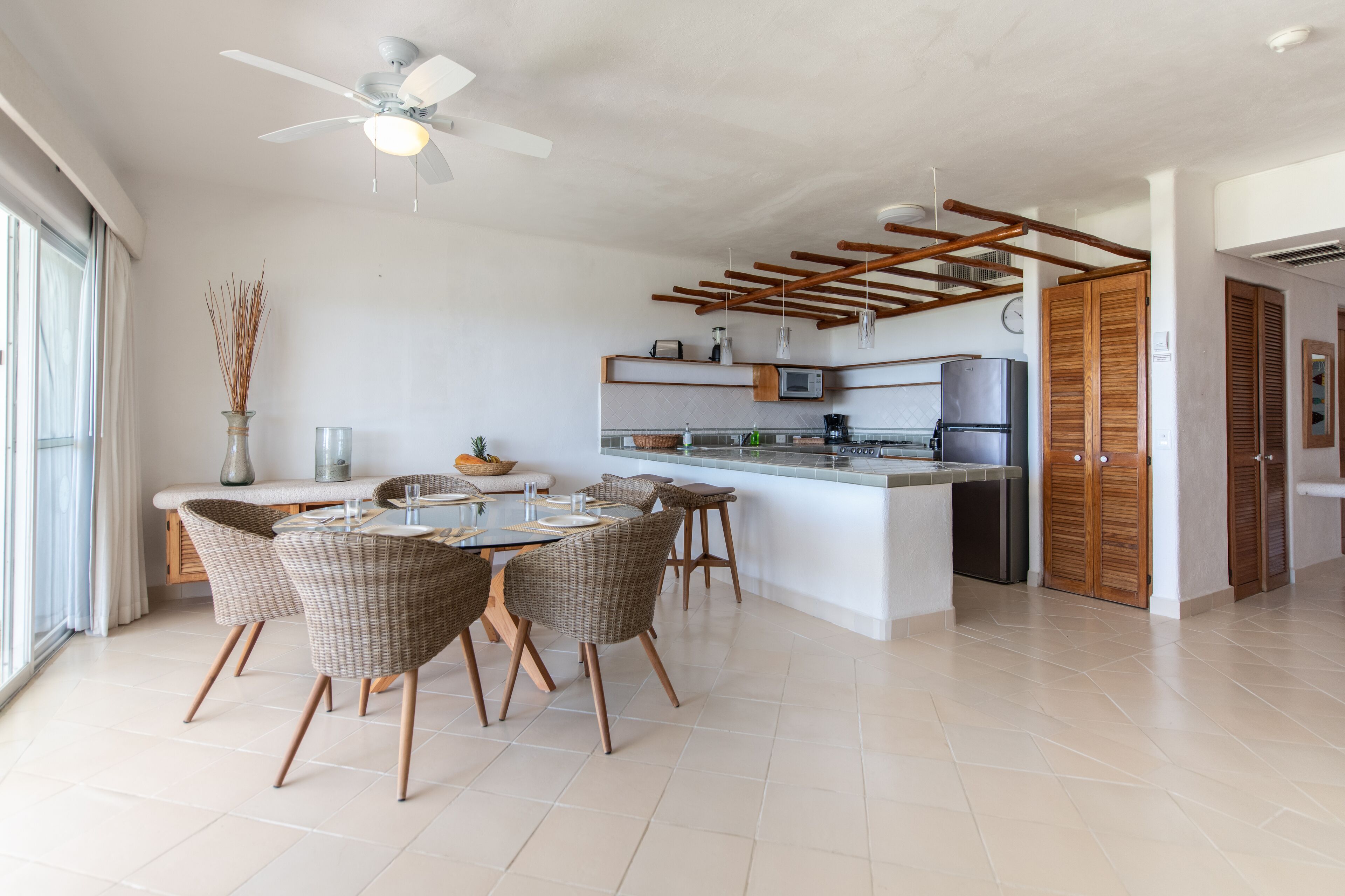 A modern kitchen with a central island, wooden ceiling beams, and a dining area with wicker chairs.