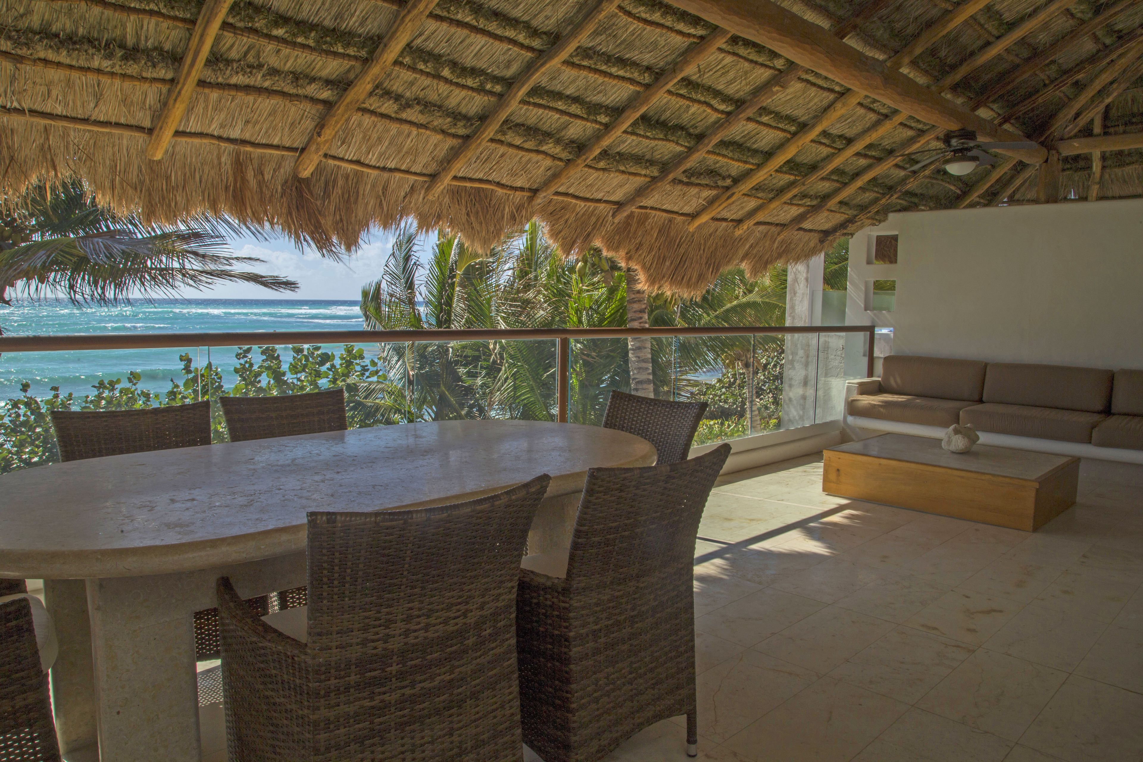 A dining area with a round table and wicker chairs, overlooking a balcony with a view of the ocean and palm trees.