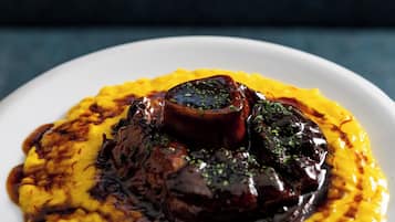 Ein weißer Teller mit gelbem Polenta und einer dunklen Soße, vermutlich Pilsoße, vor dunklem Hintergrund.