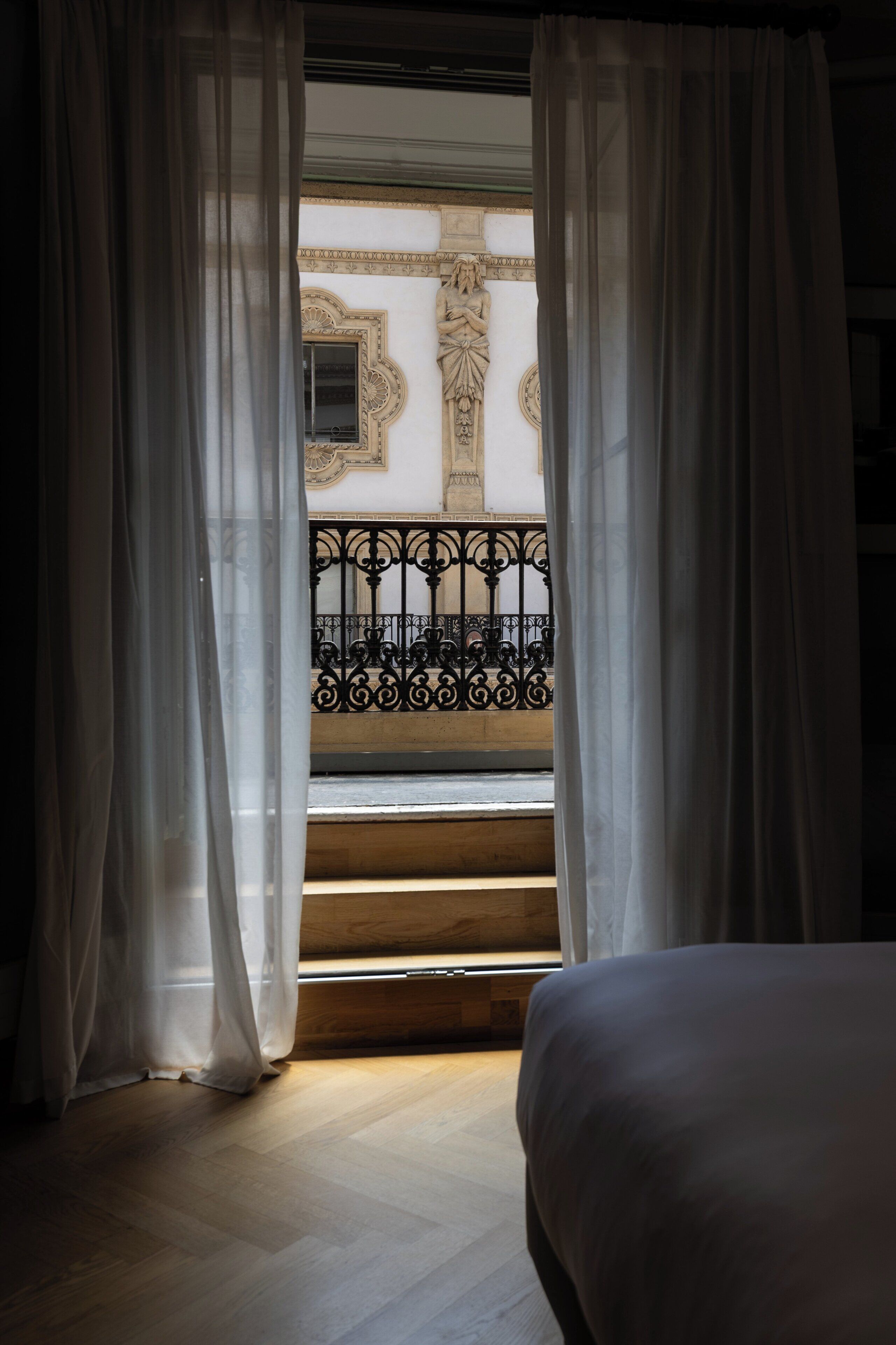 Ein Zimmer mit Bett, einem Fenster mit durchscheinenden Vorhängen und Blick auf einen Balkon mit aufwendiger Geländerarbeit.