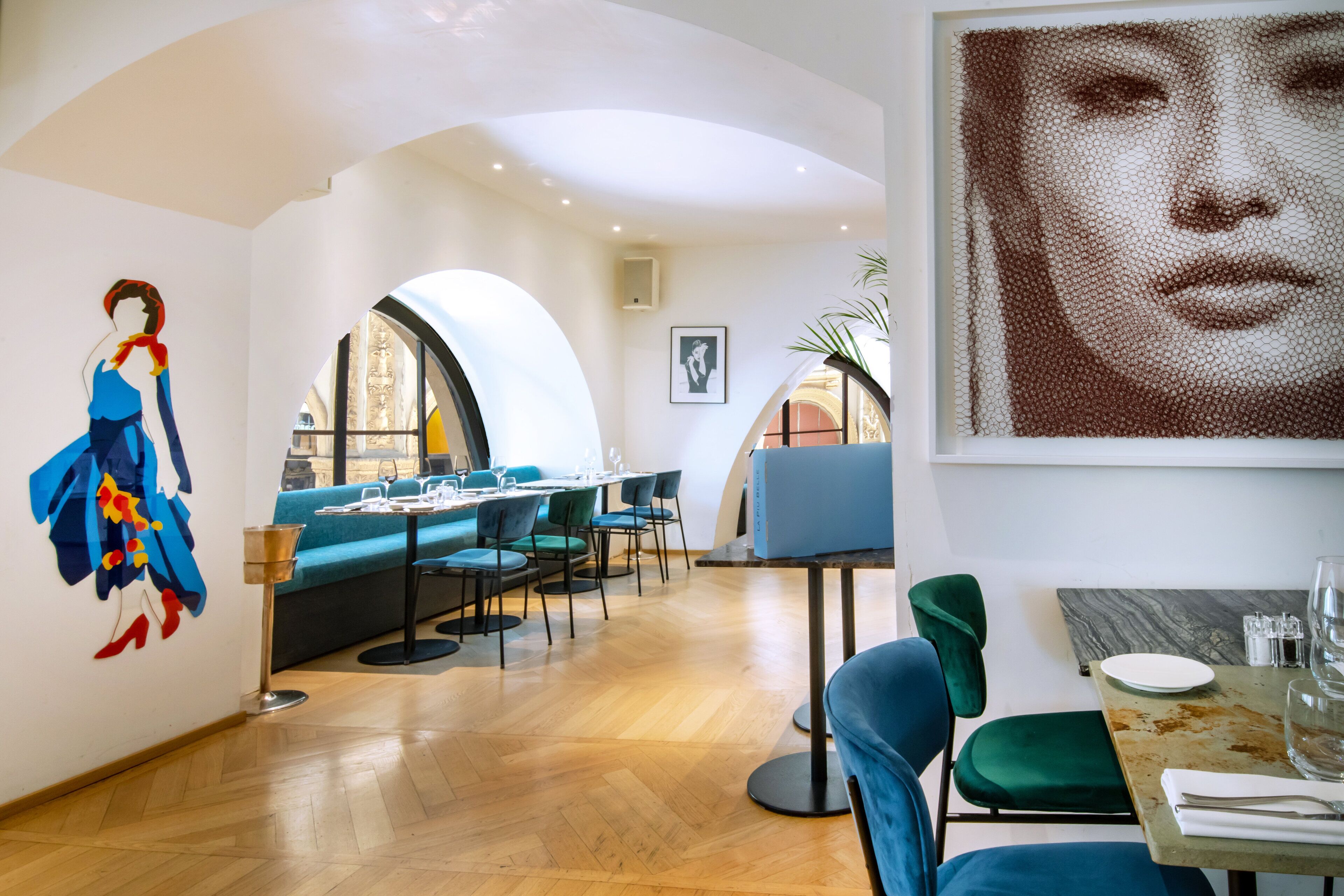 A modern dining area with a wooden floor, blue and green upholstered seating, and a large painting on the wall.