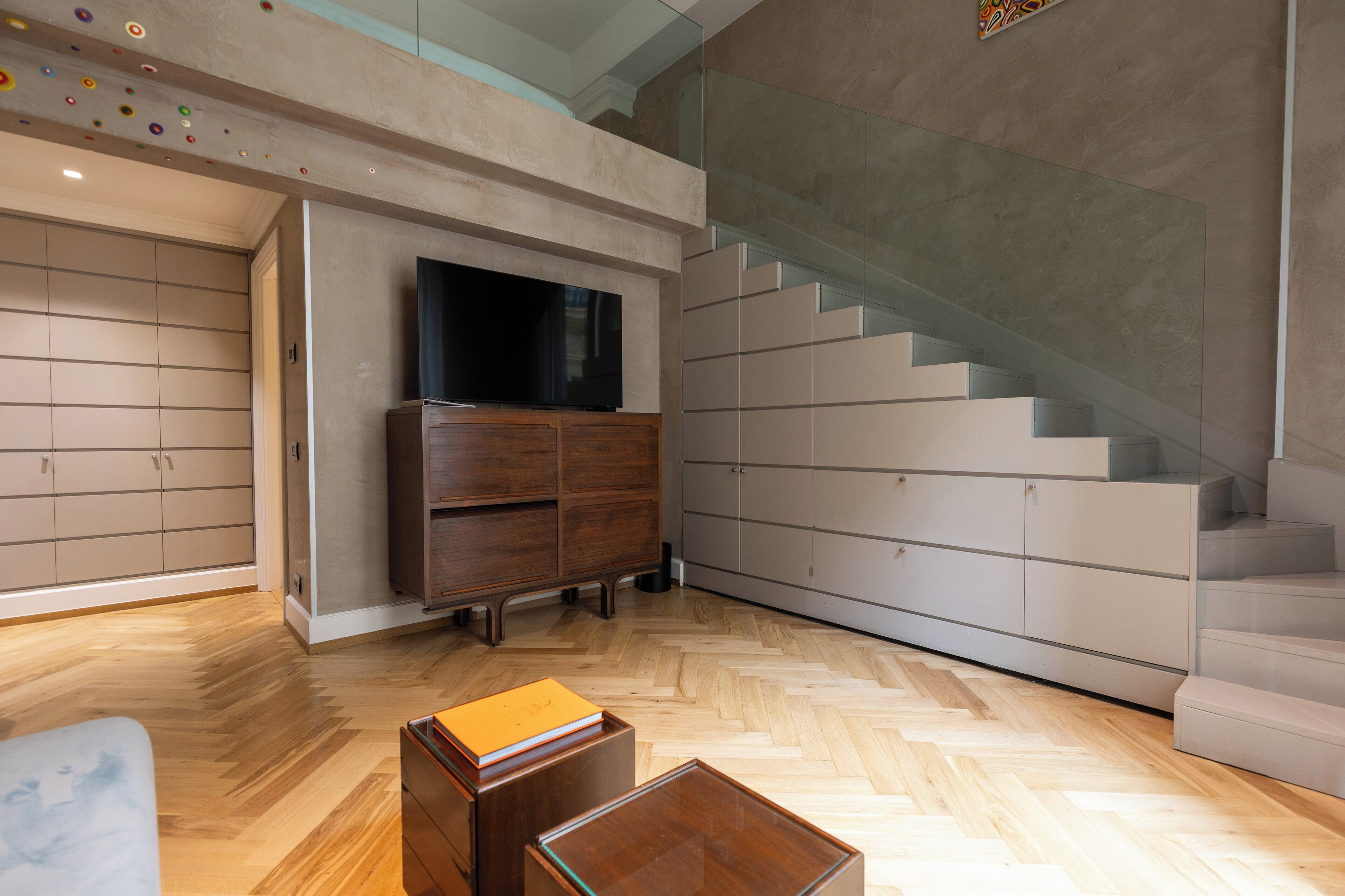 A modern interior with a herringbone wood floor, a flat-screen TV on a wooden cabinet, and a glass staircase.