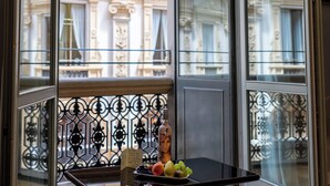 Habitación con vistas a edificios ornamentados a través de un ventanal, una pequeña mesa con frutas y una barandilla de metal.