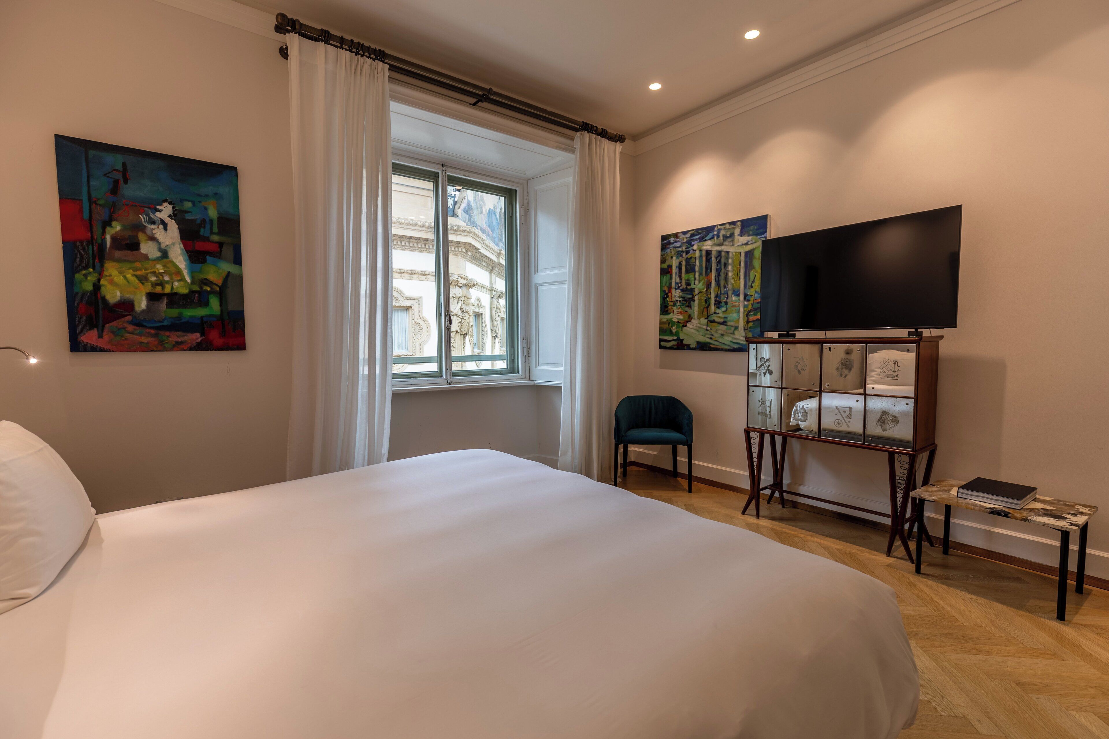 A hotel room with a large bed, a window with curtains, a television, and artwork on the walls.