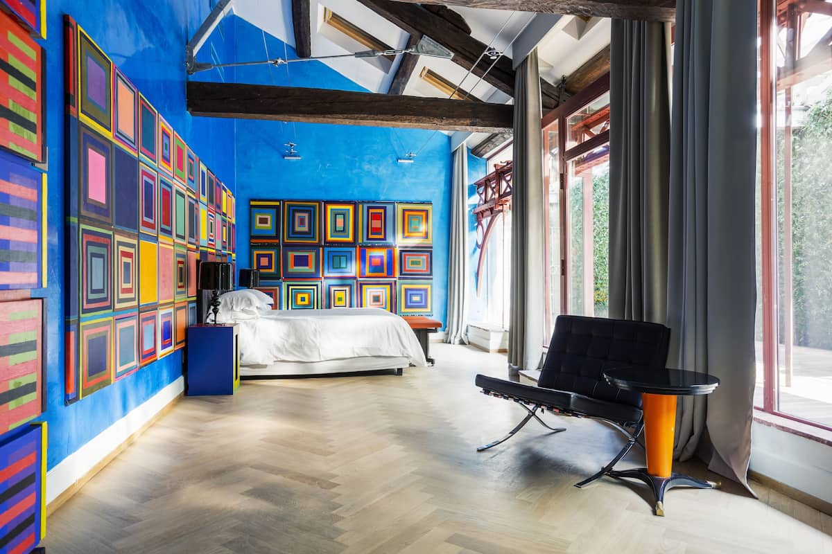 Une chambre d’hôtel moderne avec un grand lit, une chaise noire et un mur décoré d’une œuvre d’art abstraite aux couleurs vives.