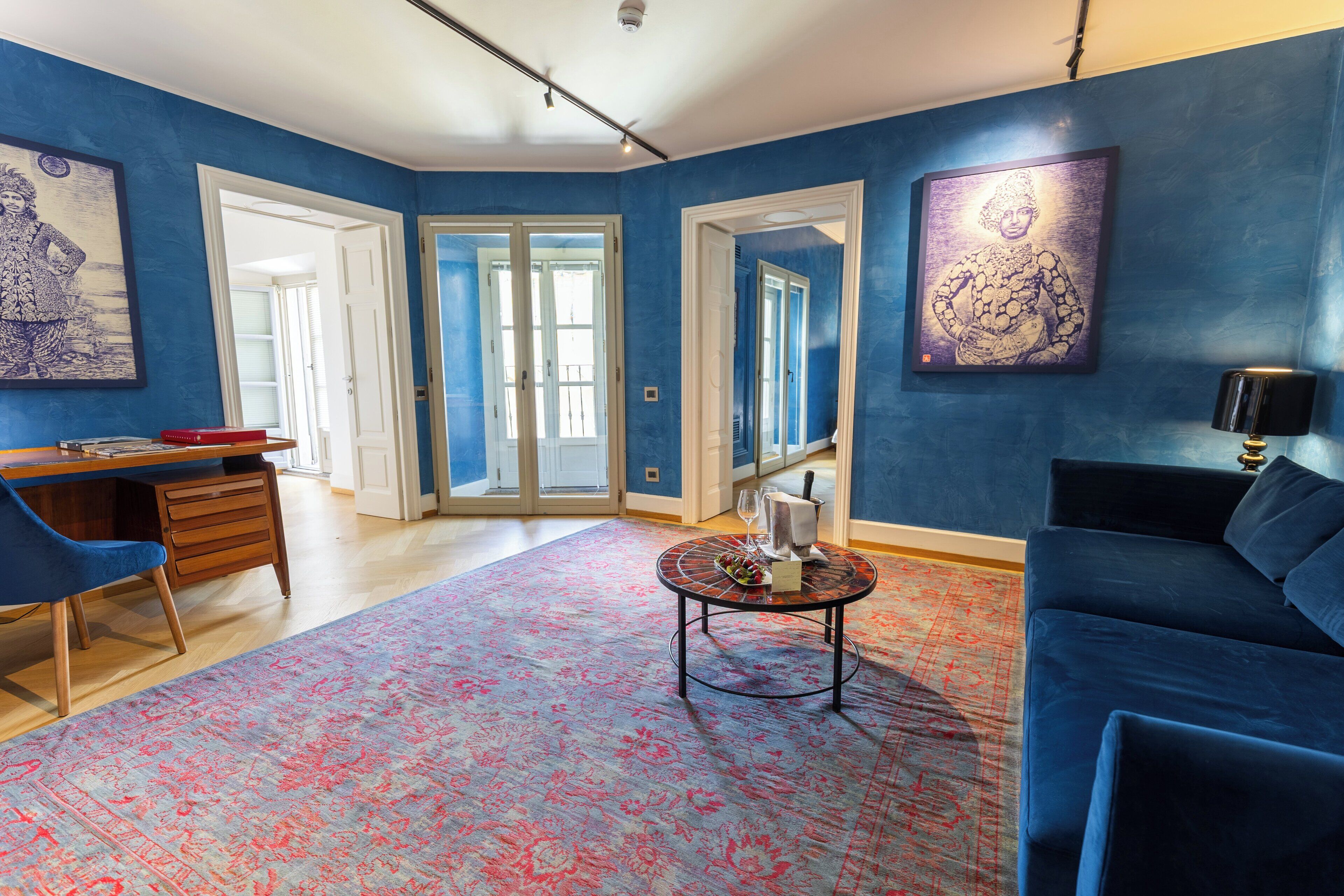 Una habitación con pared azul, un sofá azul, un escritorio con una silla, una mesa de centro y una puerta que da a otra habitación.