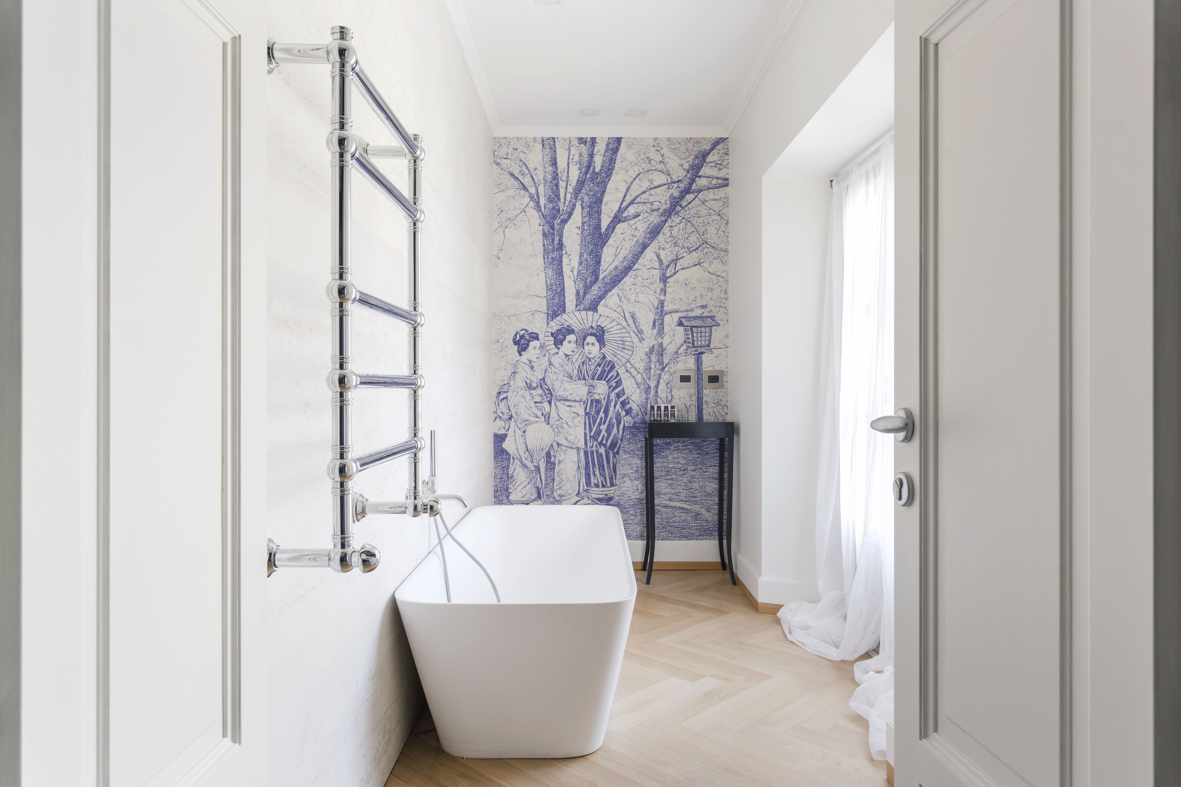 A modern bathroom with a white bathtub, a chrome towel rack, and a wall mural of a tree and figures.