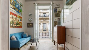 Una sala de estar moderna con un sofá azul, una mesa de centro de vidrio y un mueble de madera. Se ve un balcón con barandilla de hierro forjado a través de una puerta abierta.