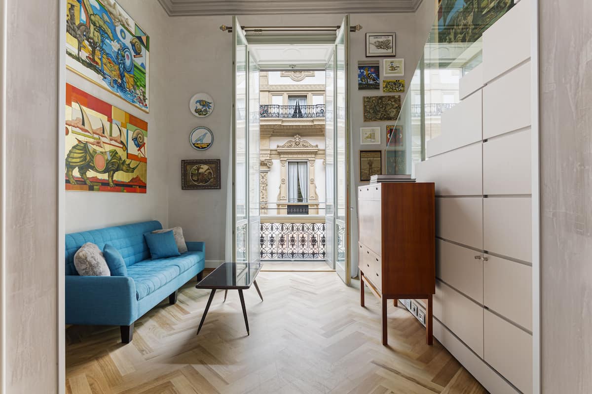 Un salon moderne avec un canapé bleu, une table basse en verre et un meuble en bois. On aperçoit un balcon avec une balustrade en fer forgé à travers une porte ouverte.