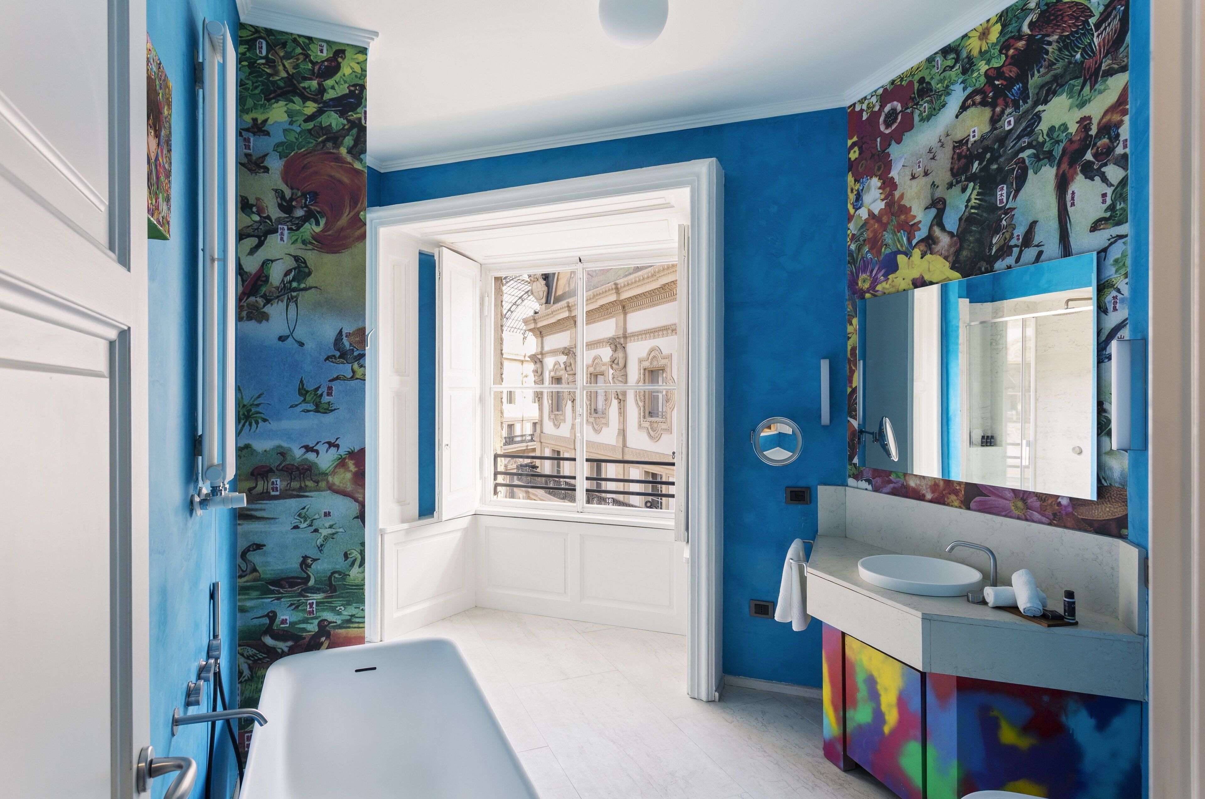 A bathroom with a large window, a white bathtub, a colorful vanity, and a mural on the wall.