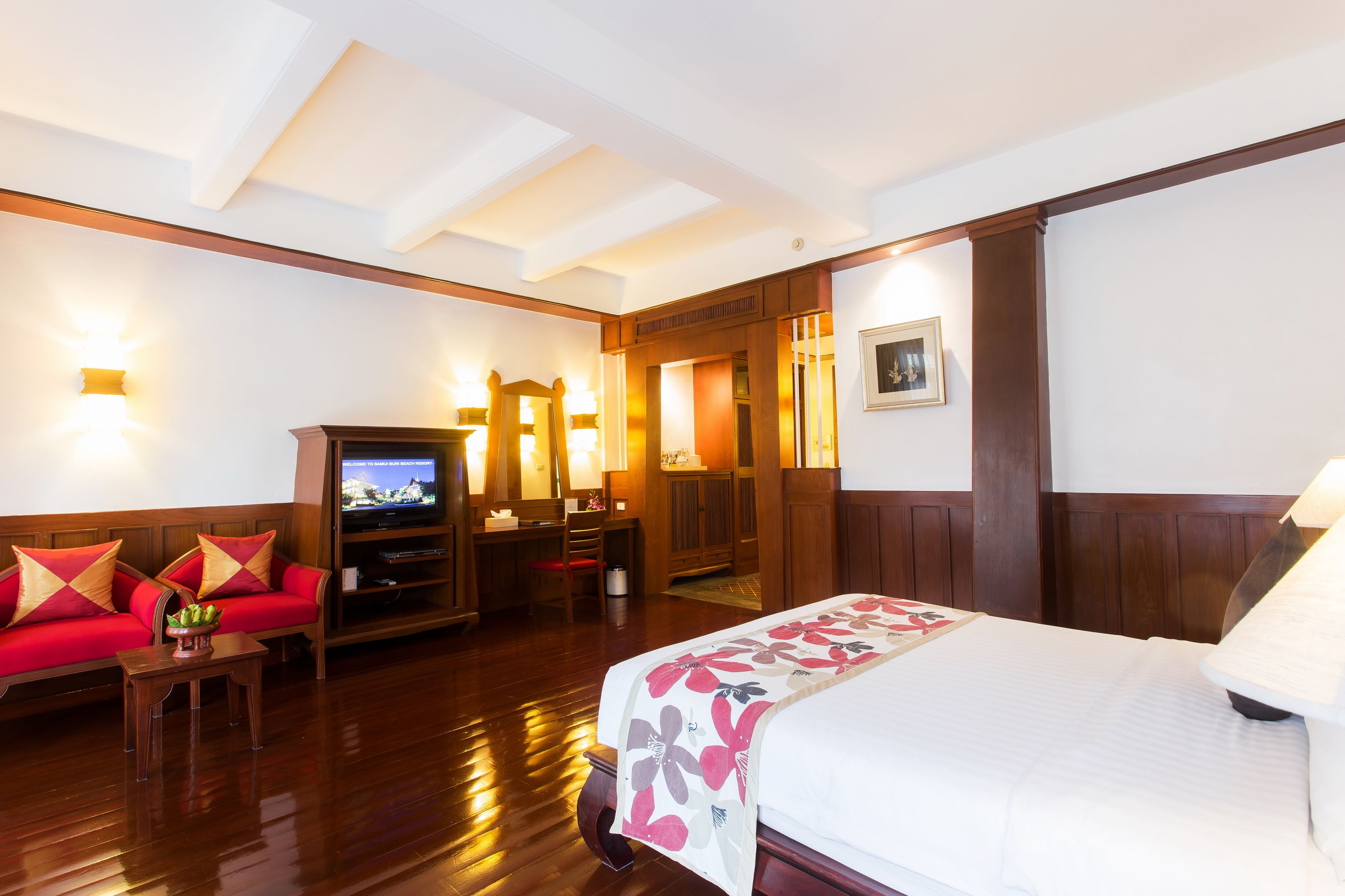 A hotel room with a wooden floor, a bed with a floral bedspread, a red sofa with cushions, a television, a desk with a chair, and a mirror.