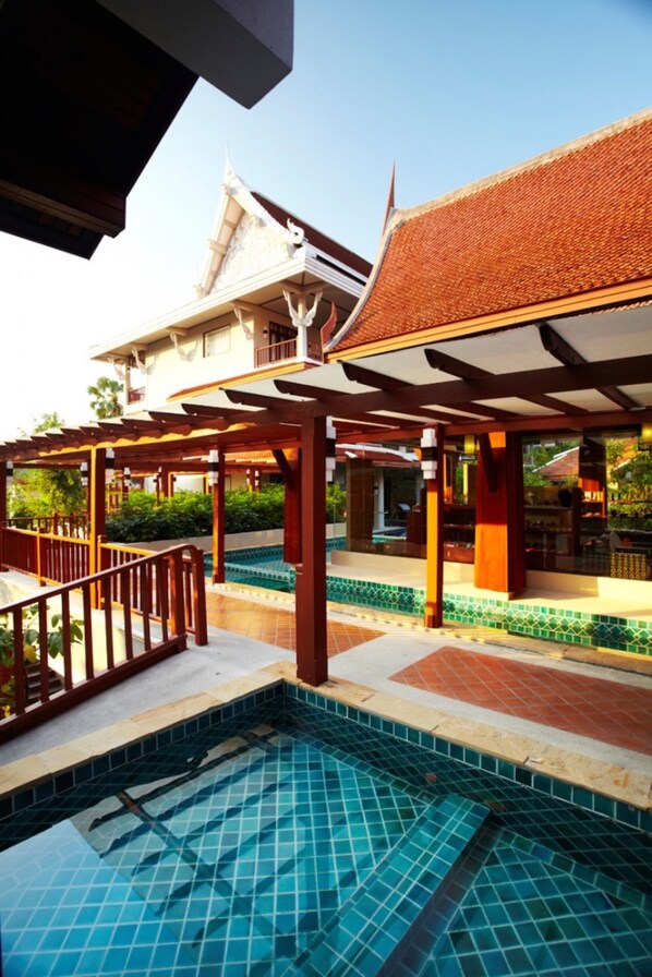 A pool area with a tiled floor, wooden pergola, and a traditional building with a tiled roof in the background.