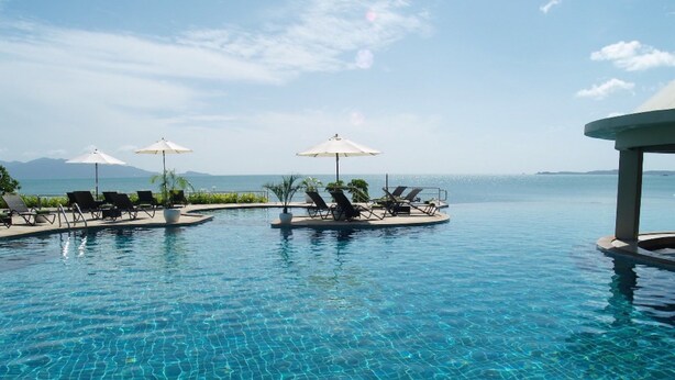 Una piscina infinita de color azul intenso con sillones de descanso y sombrillas, con vista a un mar tranquilo y montañas lejanas.
