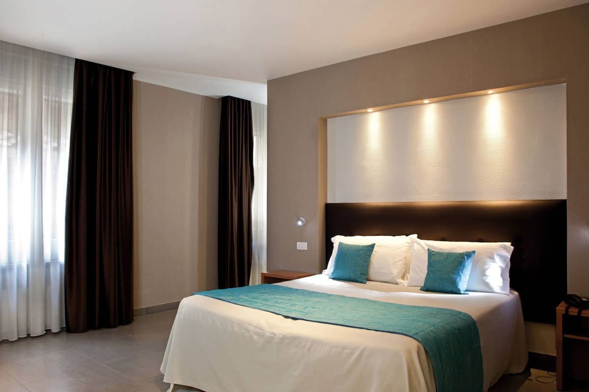 A hotel room with a bed, curtains, a headboard, and a wall-mounted light fixture.