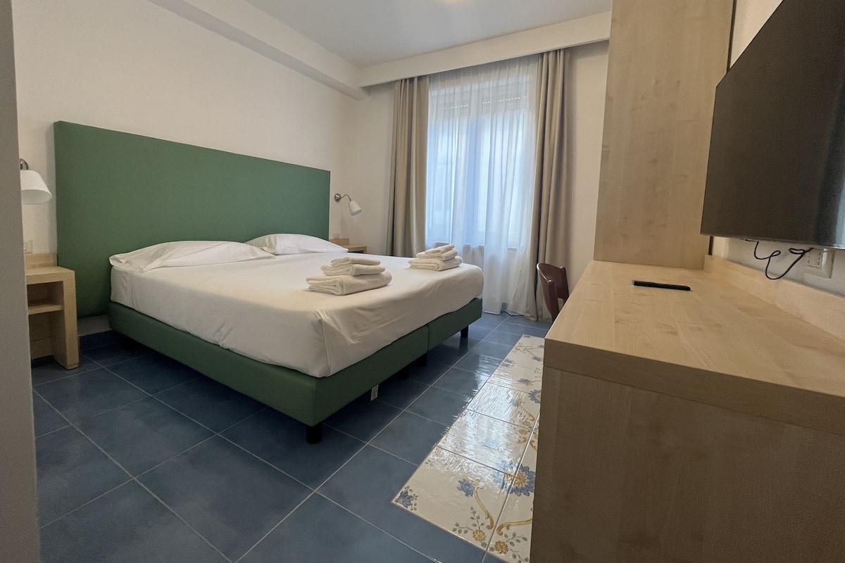 A hotel room with a green headboard, a bed with white linens, a bedside table with a lamp, a bathroom mirror, and a tiled floor.