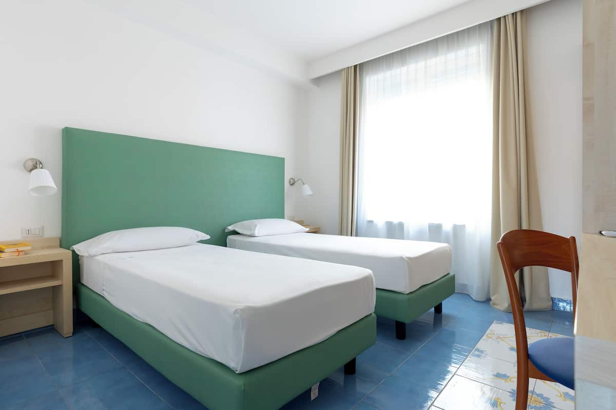 A hotel room with two single beds, a wooden chair, a nightstand, and a window with curtains.