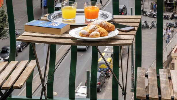 Un balcón con una mesa que sostiene un plato de cruasanes y vasos de jugo de naranja, con vistas a una calle de la ciudad con edificios históricos y un castillo.