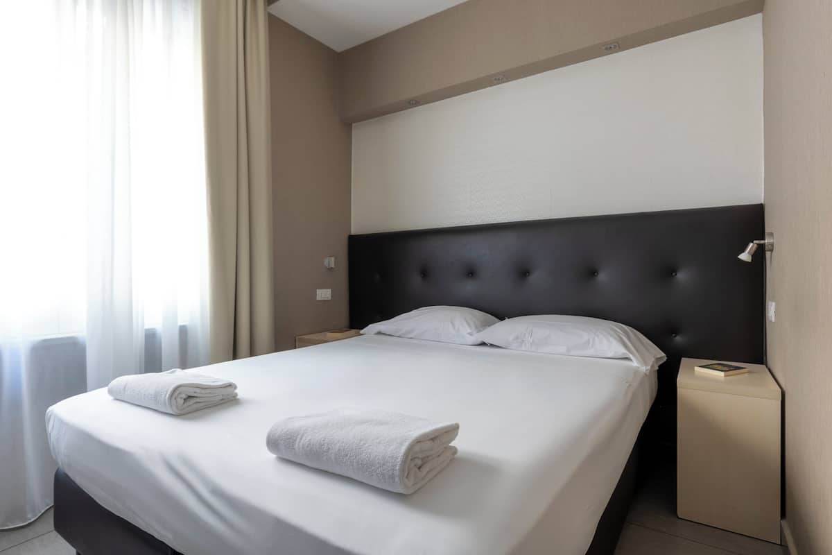A neatly made bed with white linens and two folded towels, a dark headboard, and a small bedside table with a book.