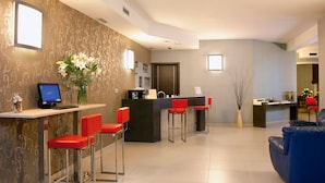 Un moderno vestíbulo de hotel con un mostrador de recepción, dos taburetes rojos, un sofá azul y un jarrón decorativo con flores blancas.
