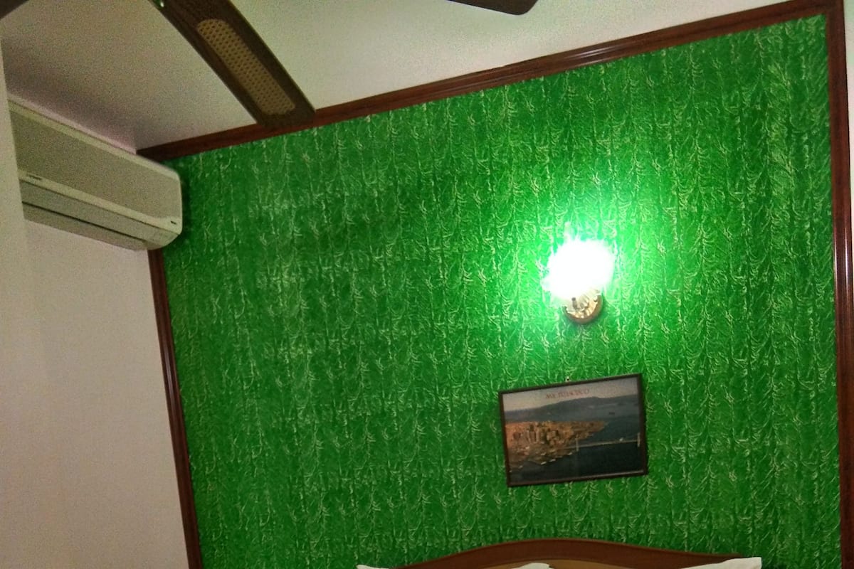 Una habitación de hotel con pared verde con textura, un cuadro enmarcado, una cama bien hecha con sábanas blancas y una manta marrón a rayas.