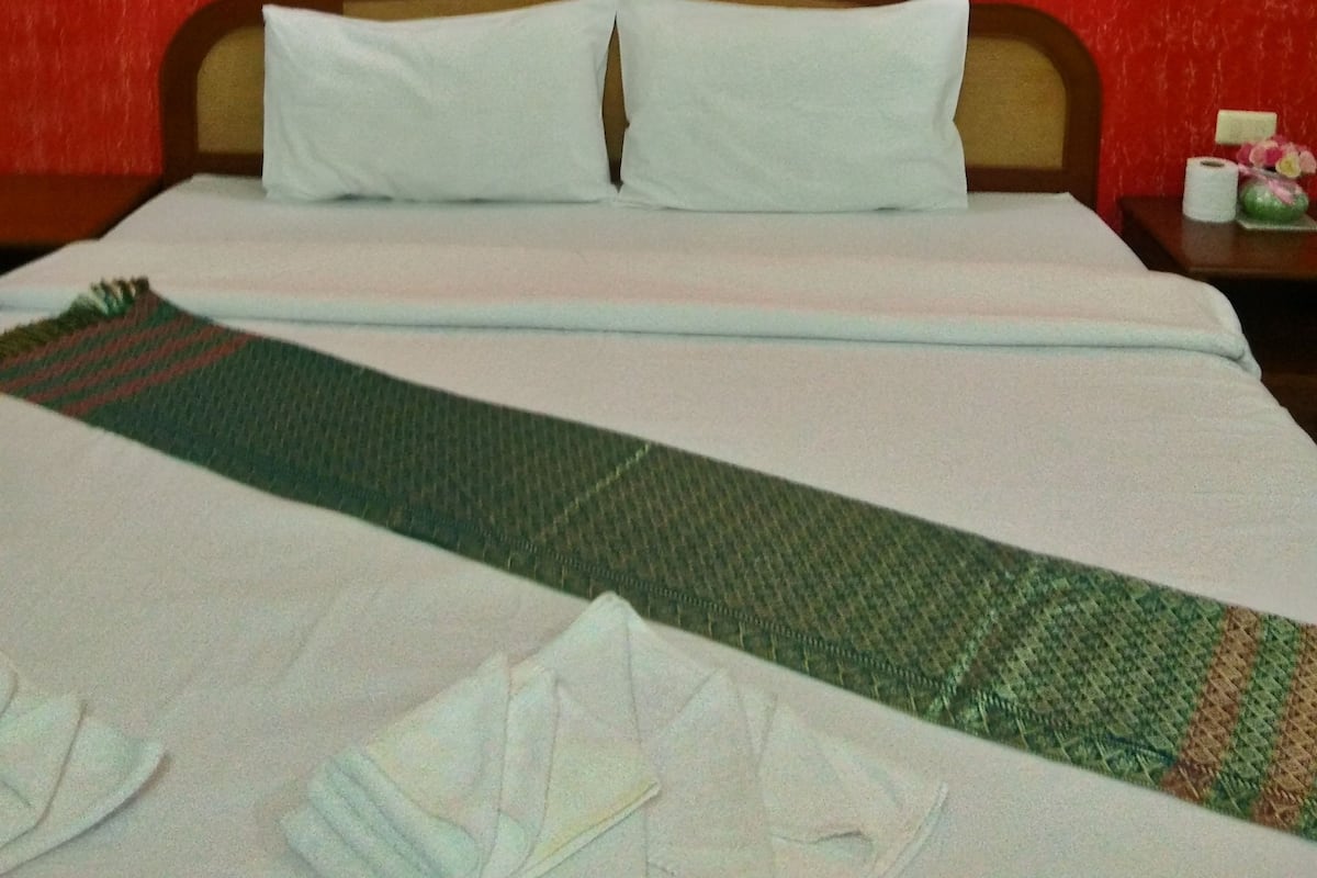 Habitación de hotel con pared roja con textura, un cuadro enmarcado, una cama bien hecha con sábanas blancas y una colcha verde estampada.