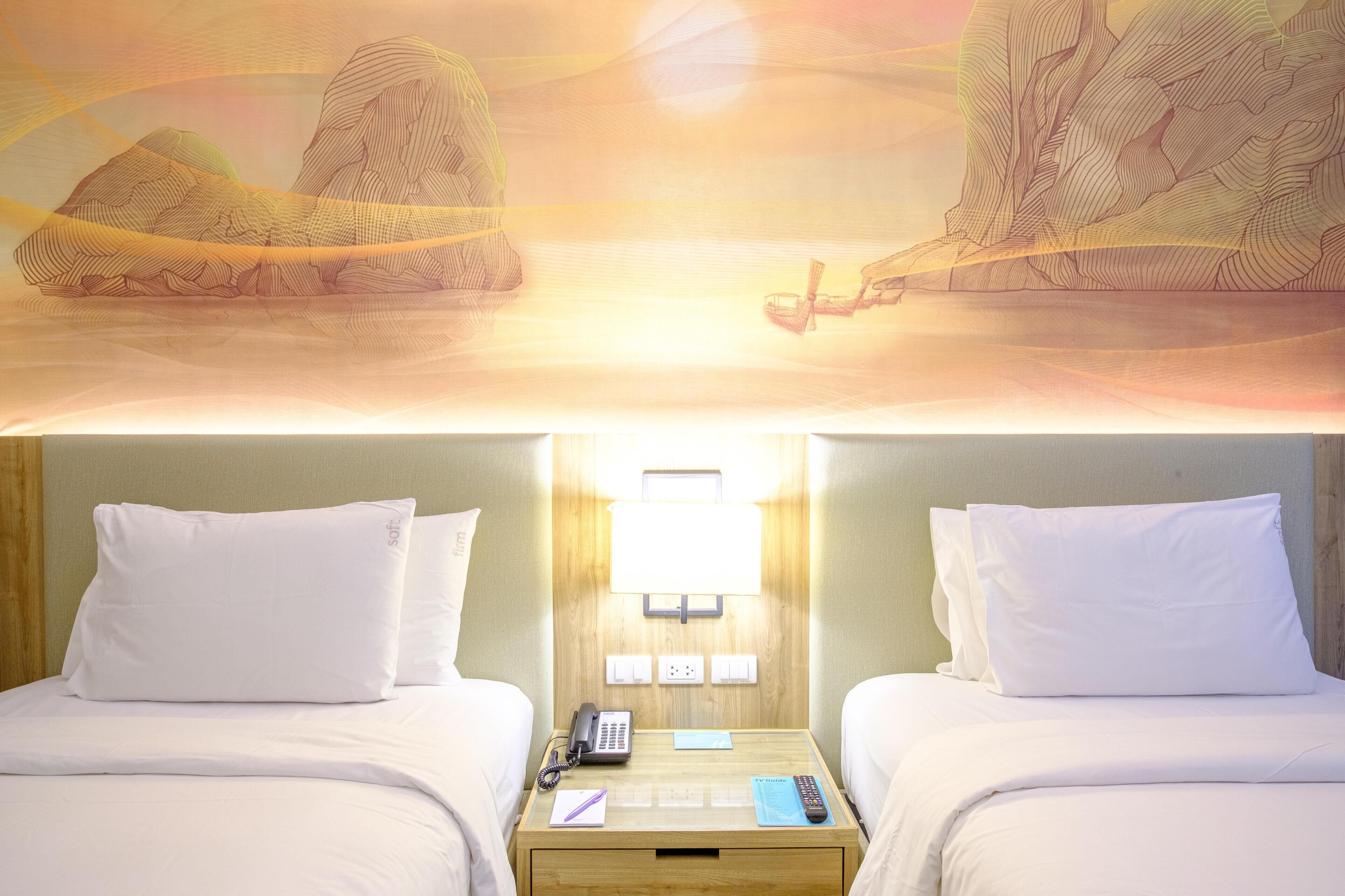 A hotel room with two beds, a bedside table with a lamp, a phone, and a remote control.