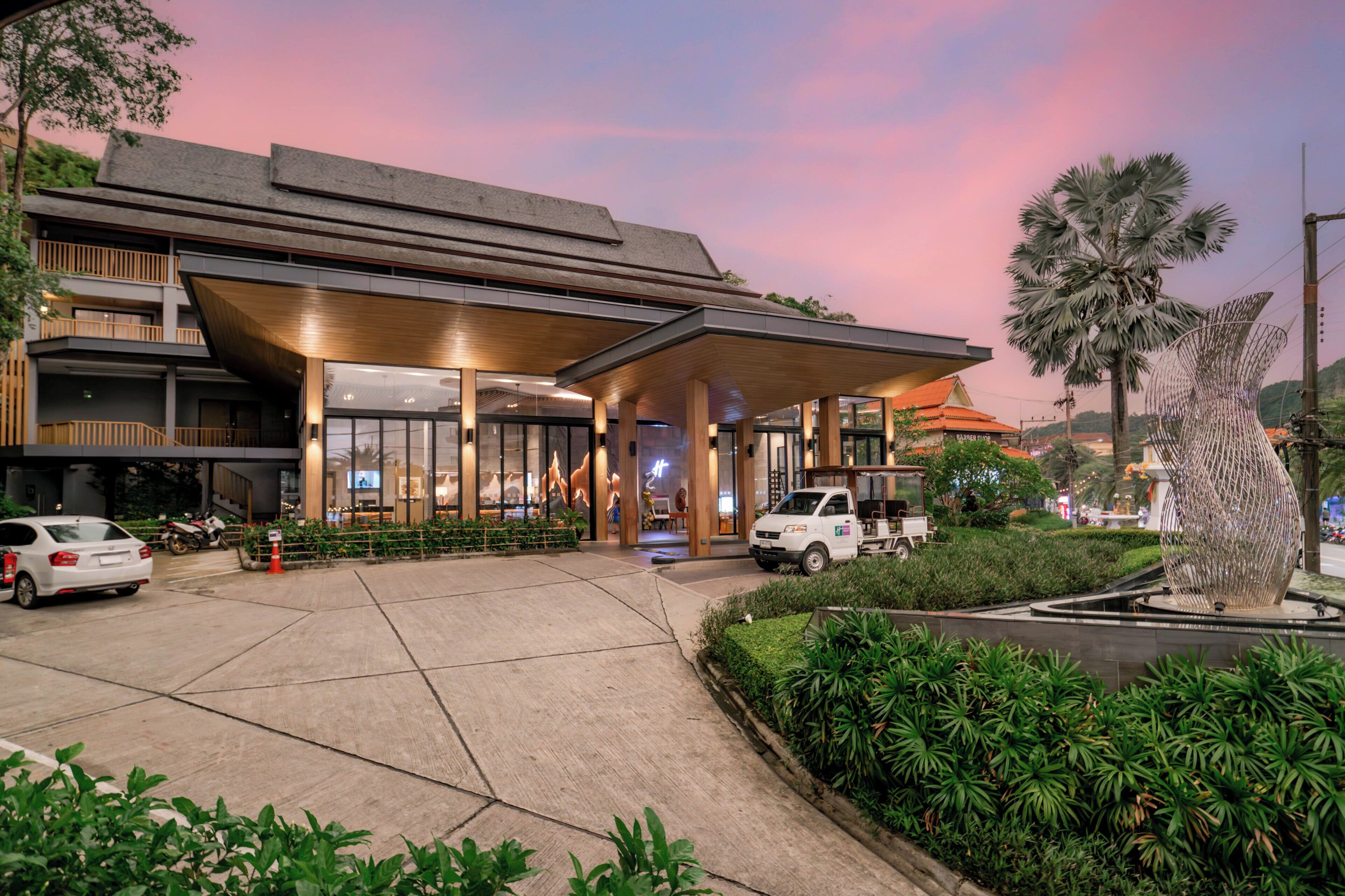 A modern building with a covered entrance, a parking area, and a landscaped garden with a palm tree.