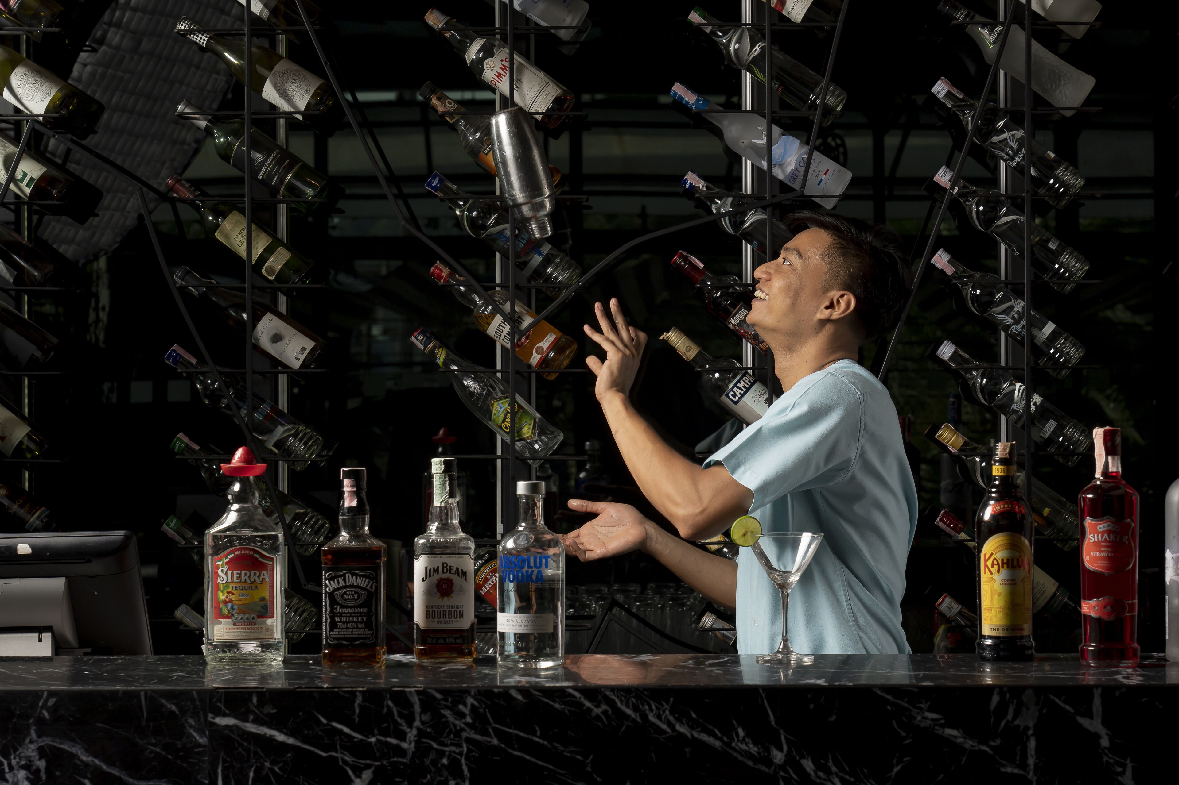 Een barman achter een bar, met verschillende drankflessen en een kassa.