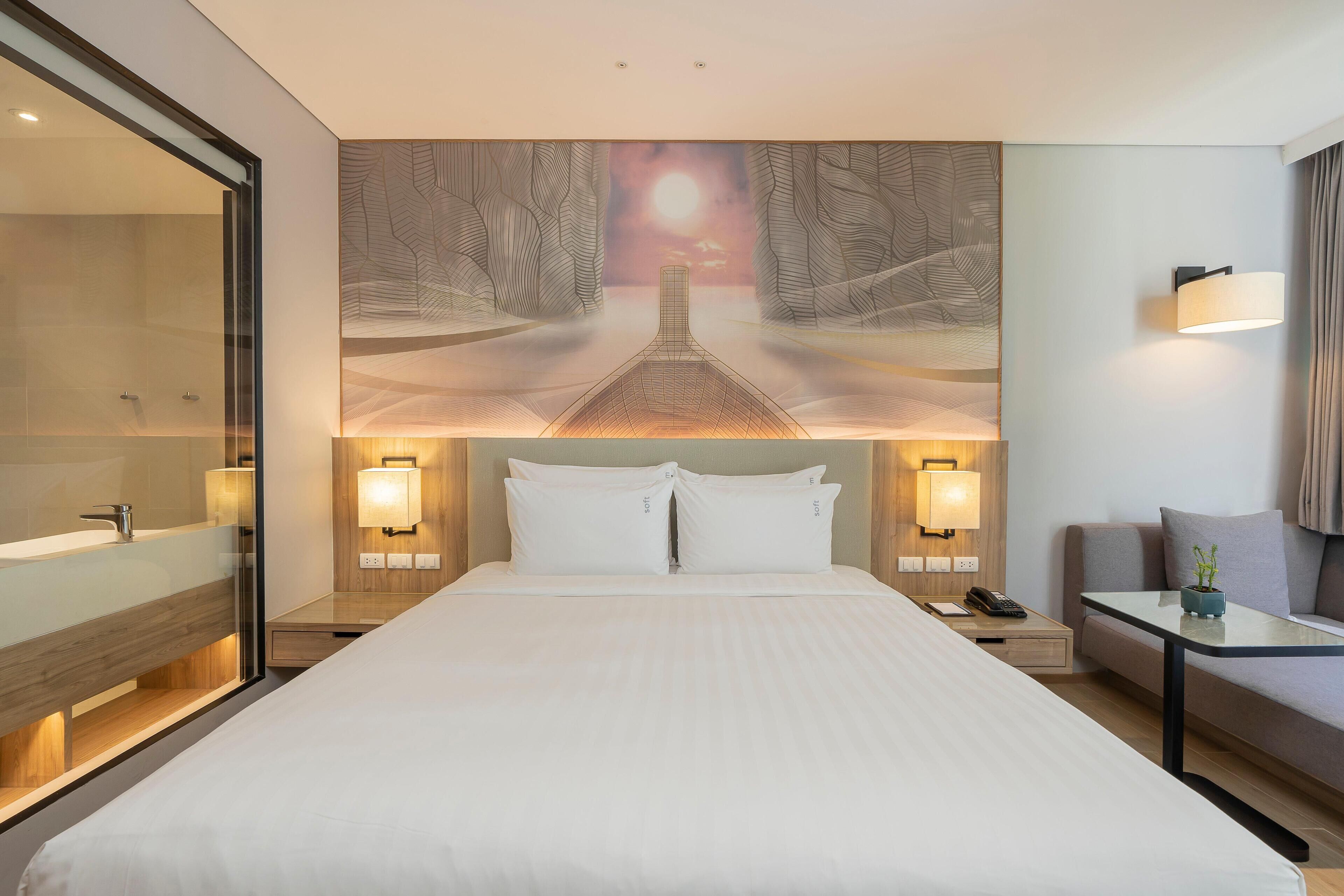 A modern hotel room with a large bed, a glass shower enclosure, a wall-mounted TV, and a seating area with a table and cushions.