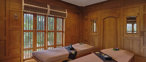 A room with wooden paneling, two massage tables, and a large window with a view of greenery.