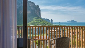 A balcony with wicker chairs and a small table, overlooking a coastal area with cliffs and a view of the sea.