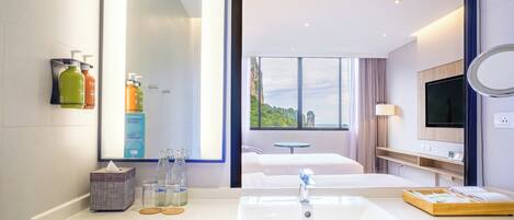 Un moderno baño de hotel con un espejo grande, un lavamanos, una bañera y una zona de ducha. Hay artículos de aseo en la encimera y una vista de un paisaje montañoso por la ventana.