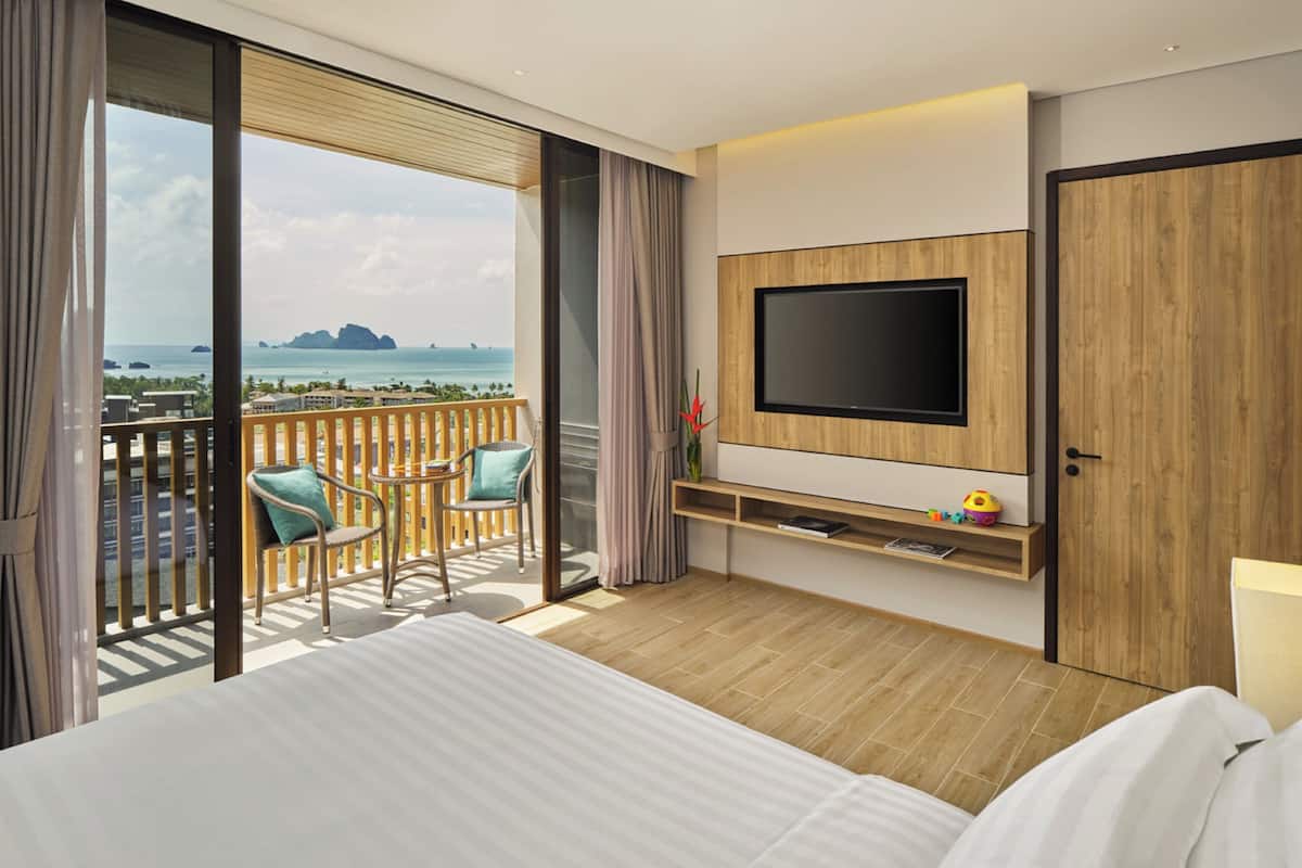 Habitación de hotel con cama, televisor montado en la pared, balcón con vistas al mar y un pequeño rincón de estar con sillas.