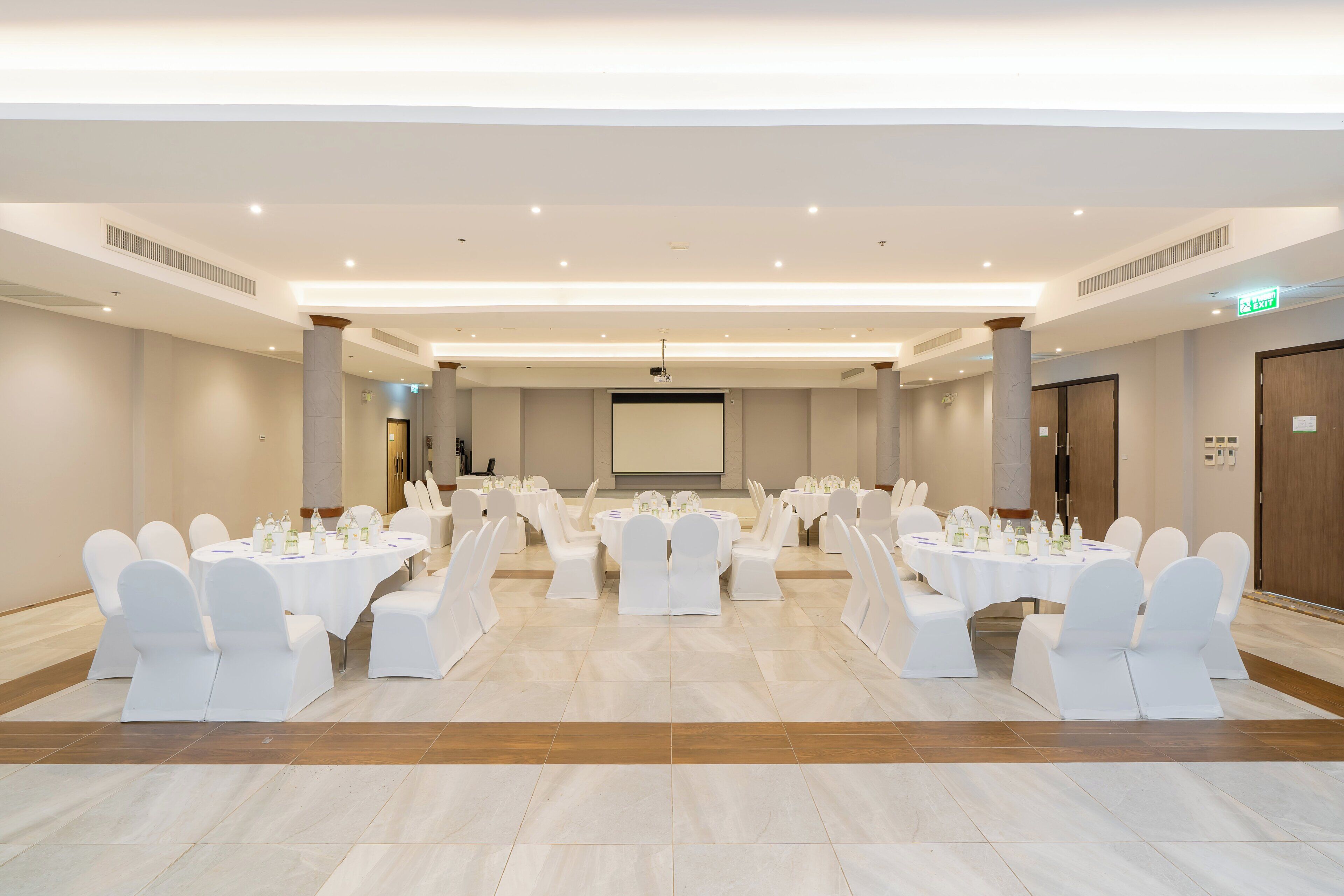Un salón de conferencias amplio con varias mesas redondas, sillas blancas y una pantalla de proyector al frente.