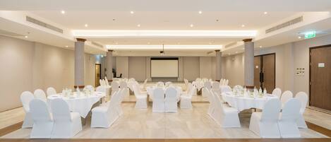 Un salón de conferencias amplio con varias mesas redondas, sillas blancas y una pantalla de proyector al frente.