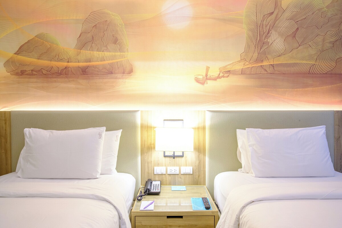 Une chambre d’hôtel avec deux lits, une table de chevet avec un téléphone et une télécommande, et un luminaire fixé au mur.
