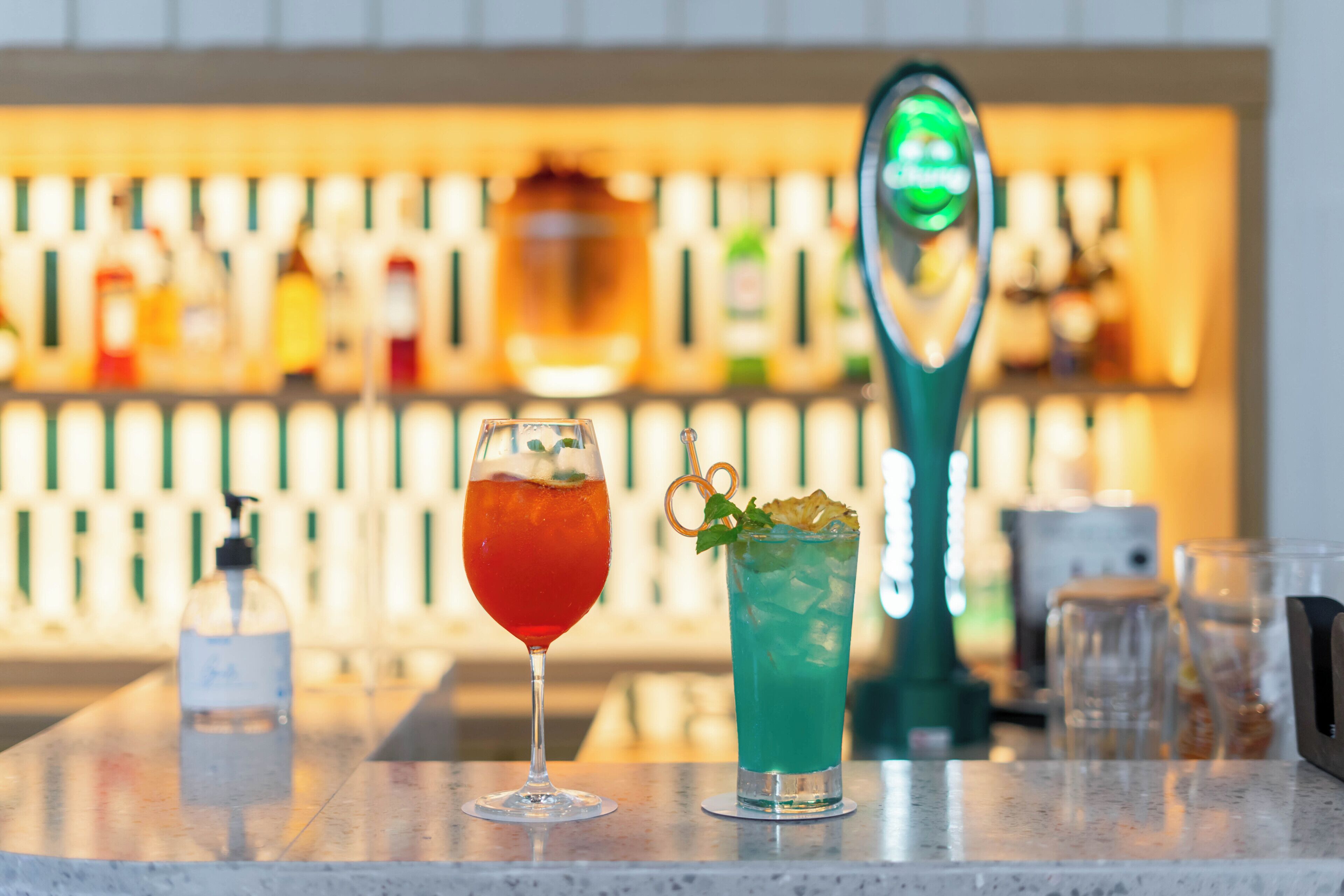 Um balcão de bar com duas bebidas, um frasco de álcool em gel e uma ferramenta de bar verde.