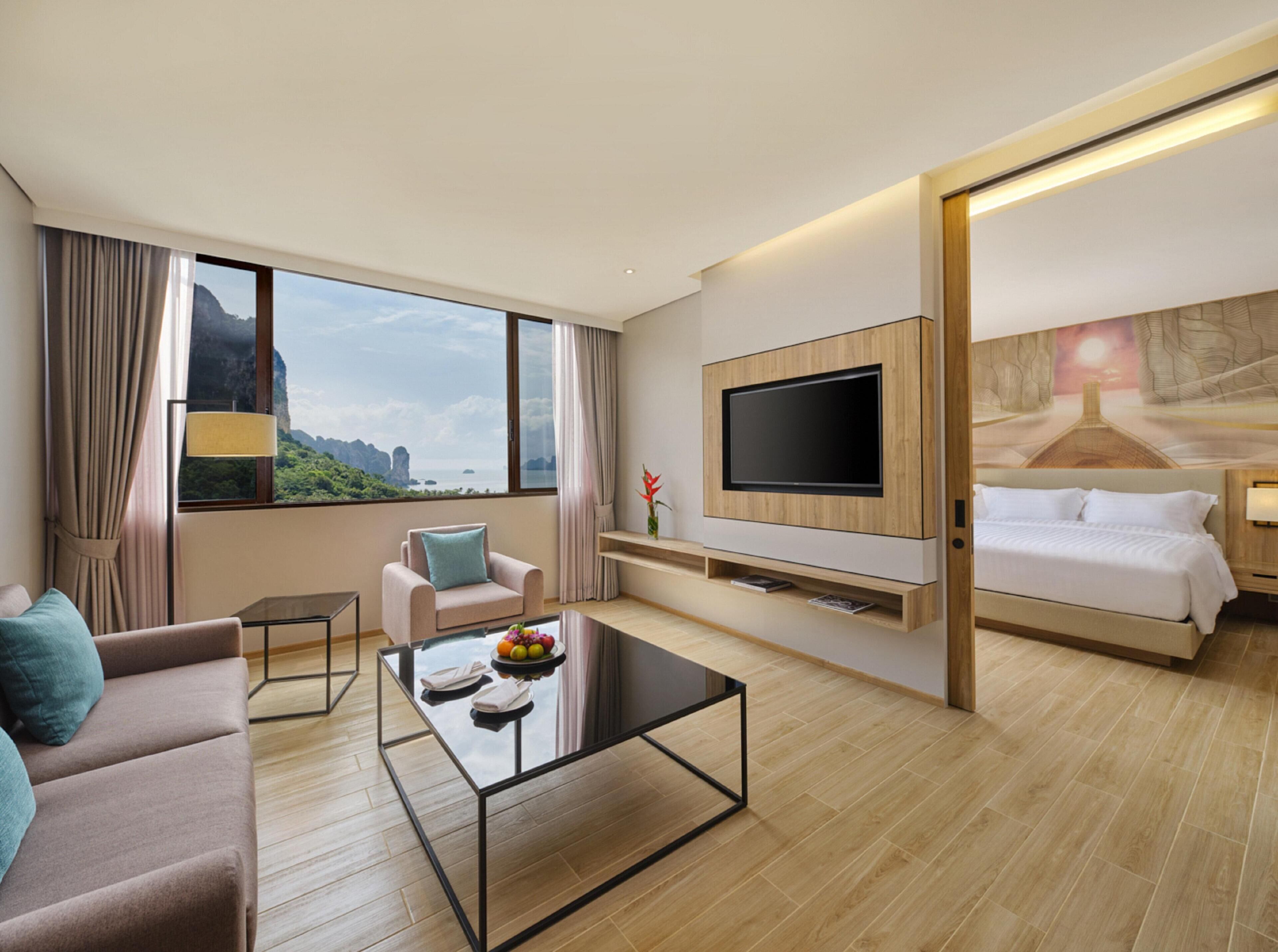 A modern hotel room with a large window, a flat-screen TV, a sofa, a coffee table, and a bed.