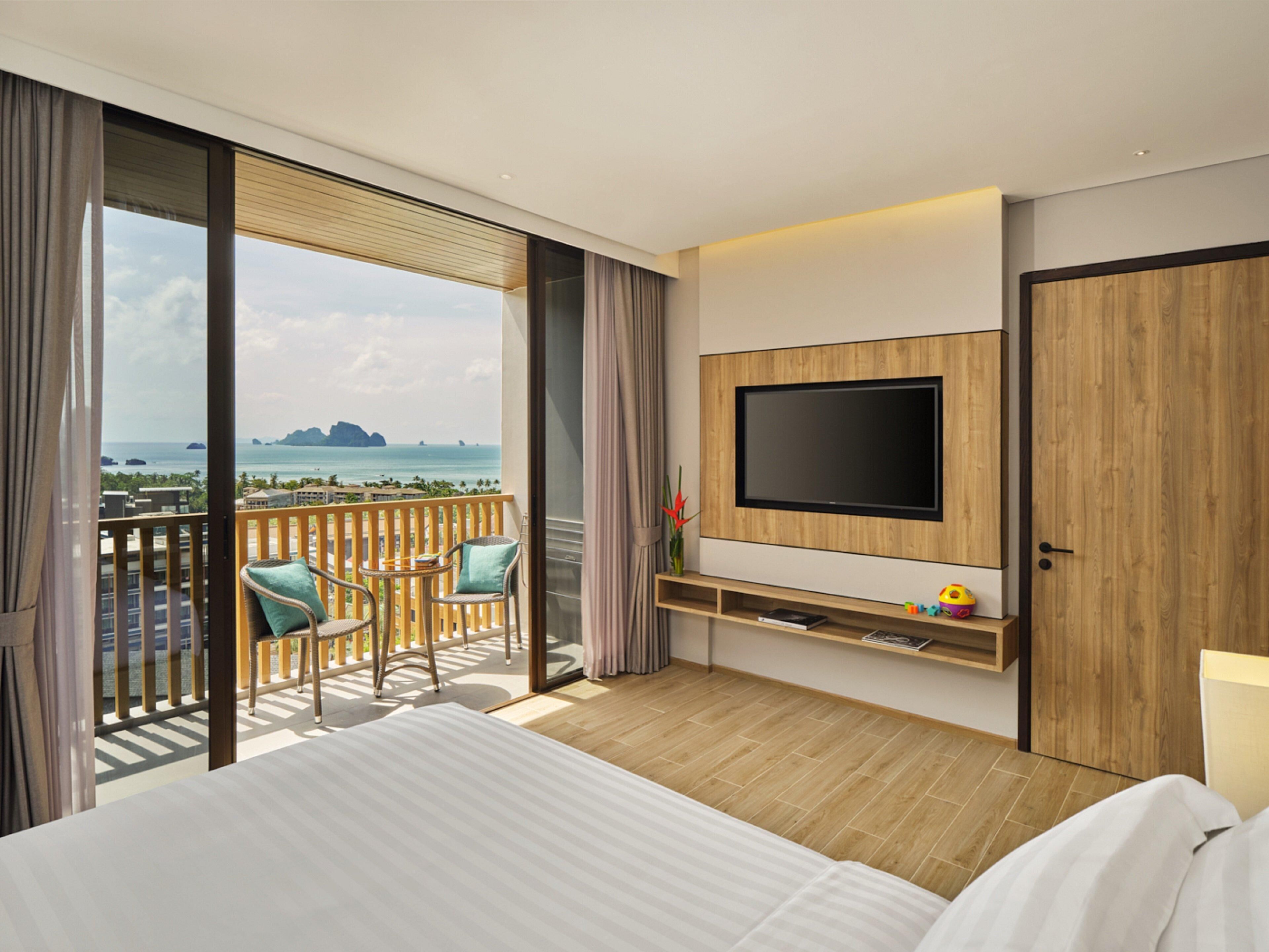 A hotel room with a bed, a TV mounted on a wooden panel, a balcony with chairs, and a view of the sea.