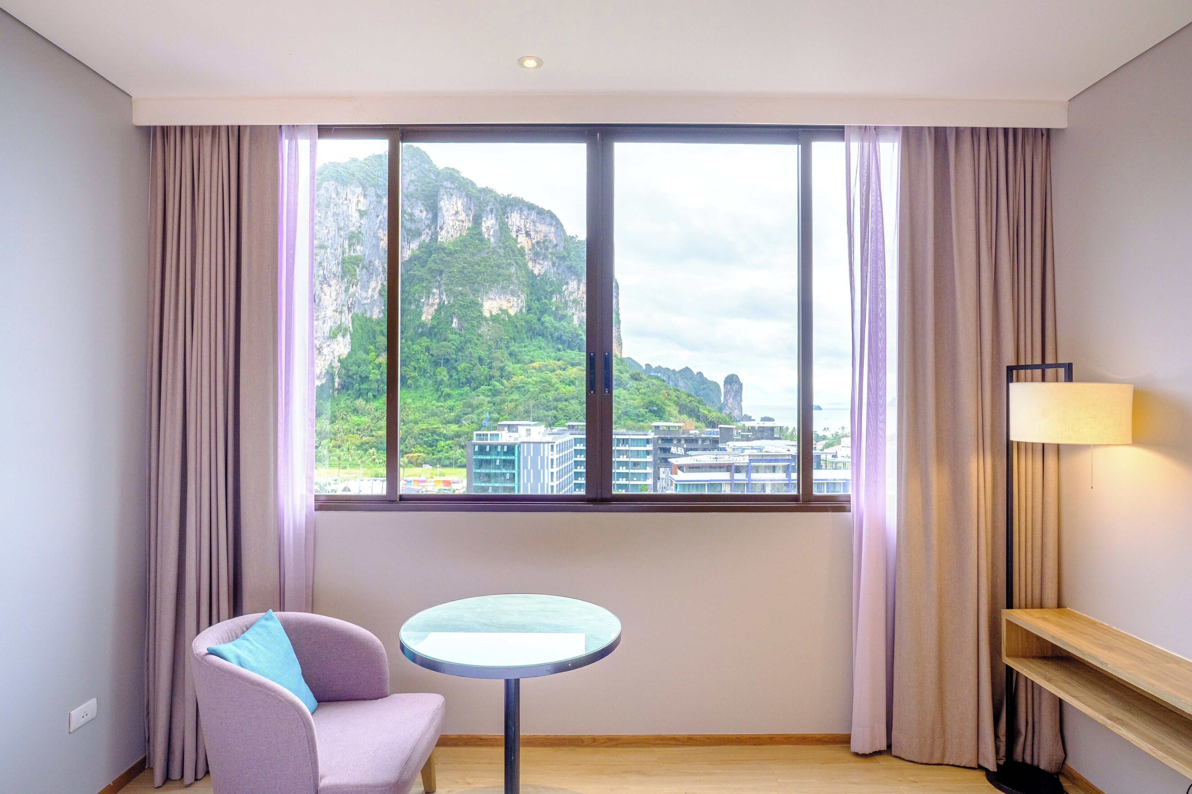 A hotel room with a large window, a round table, a chair, and a lamp.
