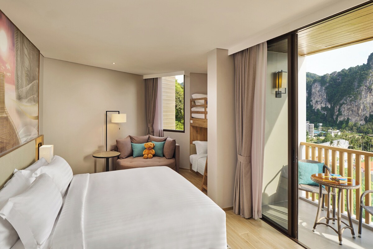 Une chambre d’hôtel avec un grand lit, un bureau, une chaise, un balcon avec vue sur les montagnes et une fenêtre avec des rideaux.