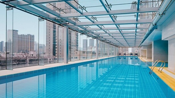 A modern indoor swimming pool with clear glass walls, a blue tiled floor, and a view of city buildings. - Sheraton Changsha Hotel (Changsha)