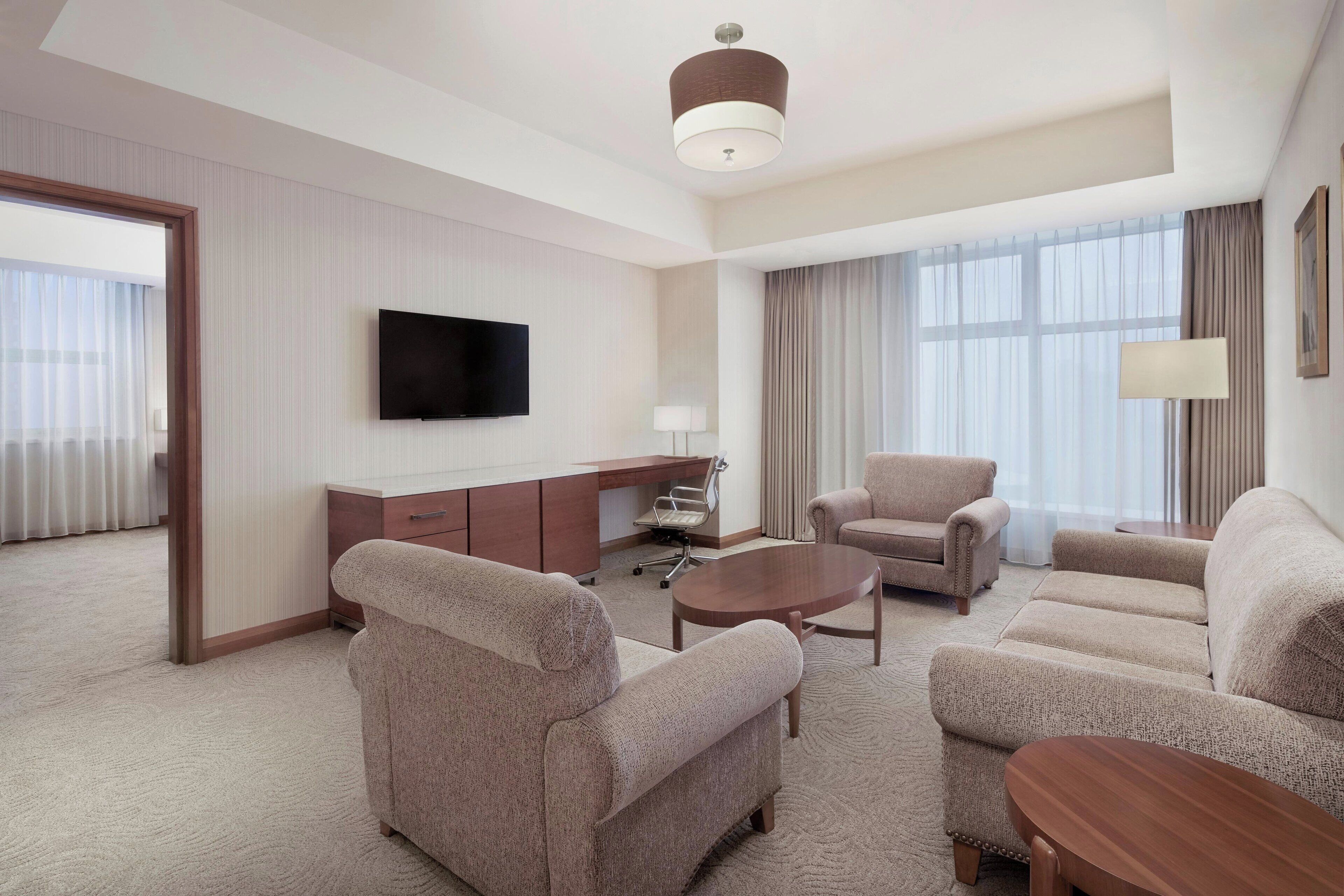 A modern hotel room with a flat-screen TV mounted on the wall, a desk with a chair, a sofa, and a coffee table.