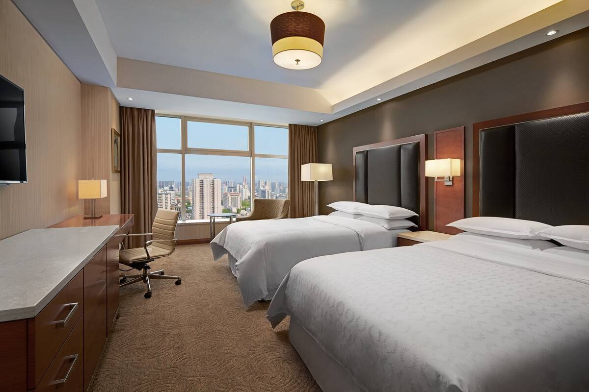 A hotel room with two beds, a desk, and a chair. There is a large window with a city view.