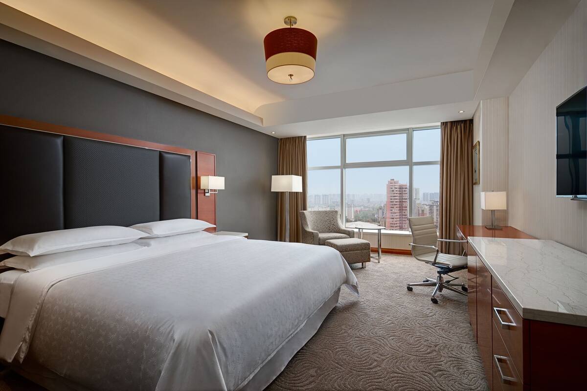 A hotel room with a large bed, two chairs, a desk, and a view of the city.
