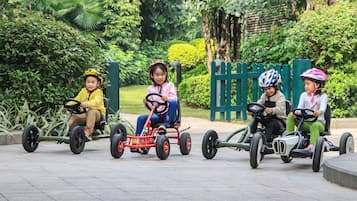 Cuatro niños conduciendo karts en un parque con follaje y una cerca verde al fondo.