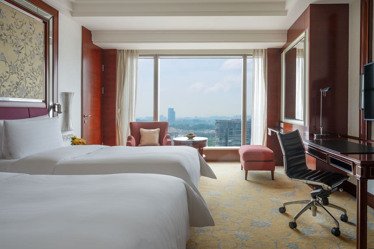 A hotel room with two beds, a desk, a chair, a TV, and a large window with a city view.