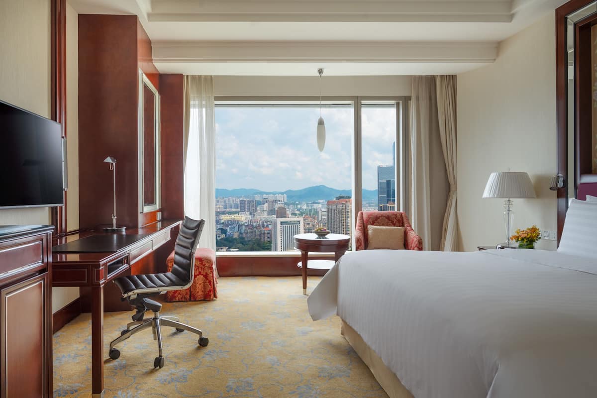 A hotel room with a large bed, a desk with a chair, a television, and a view of the cityscape.