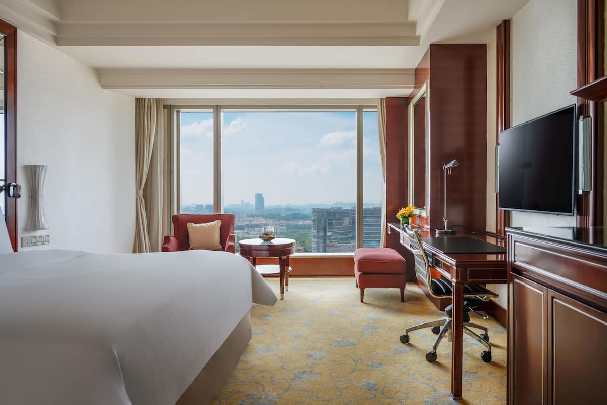 A hotel room with a large bed, a desk with a chair, a television, and a view of the cityscape through large windows.
