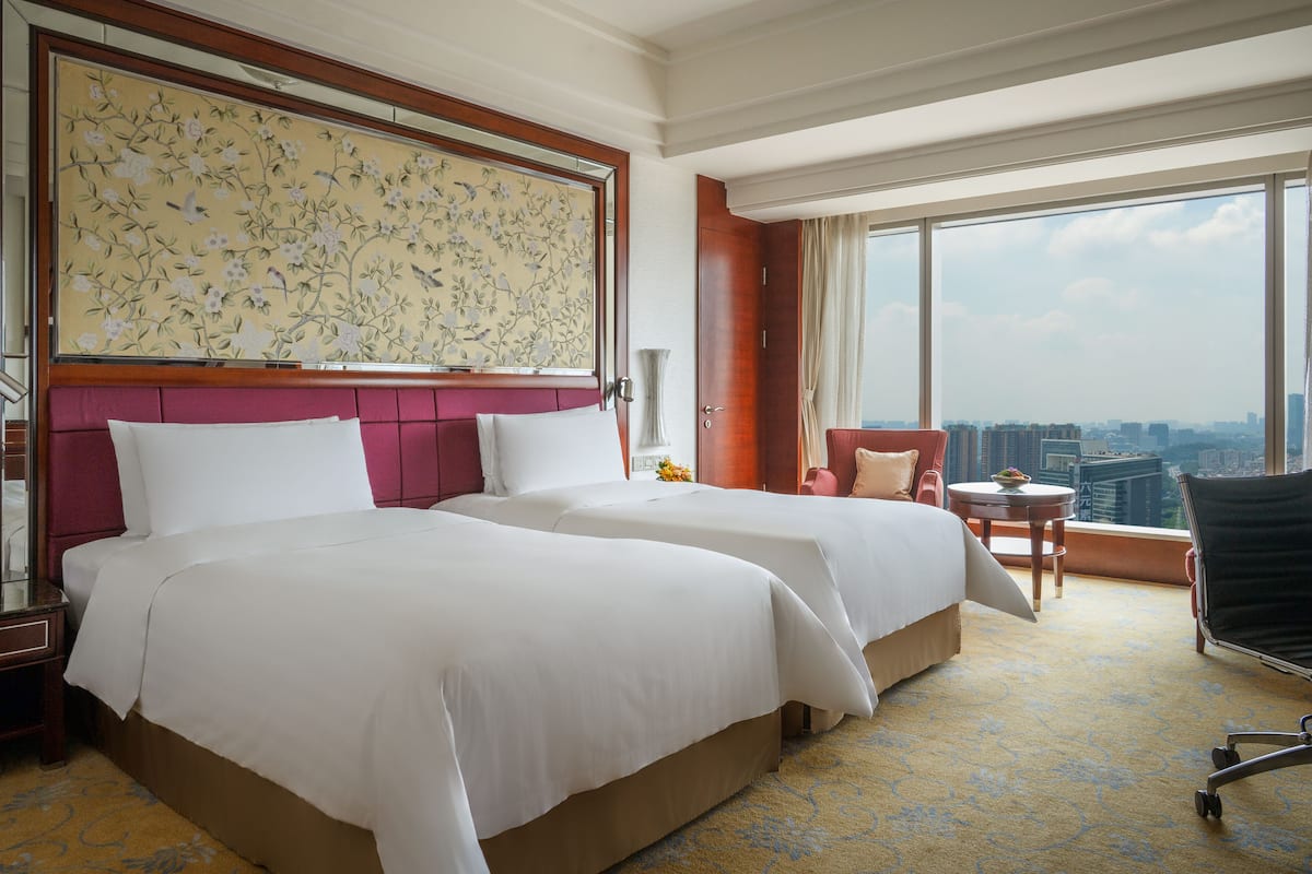 A hotel room with two beds, a large floral artwork on the wall, a city view through the window, and a small round table with a chair.